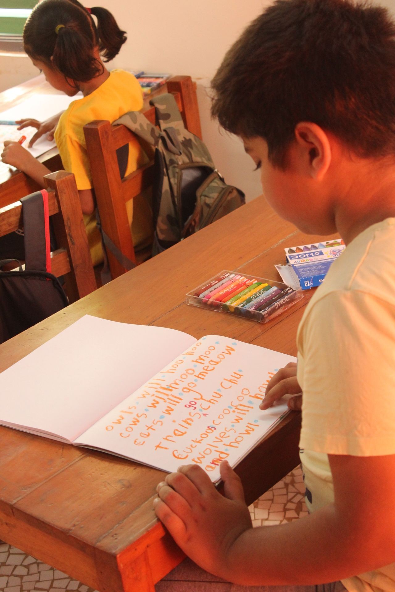 Children sitting at a desk, one reading a notebook with colorful writing in a classroom setting.