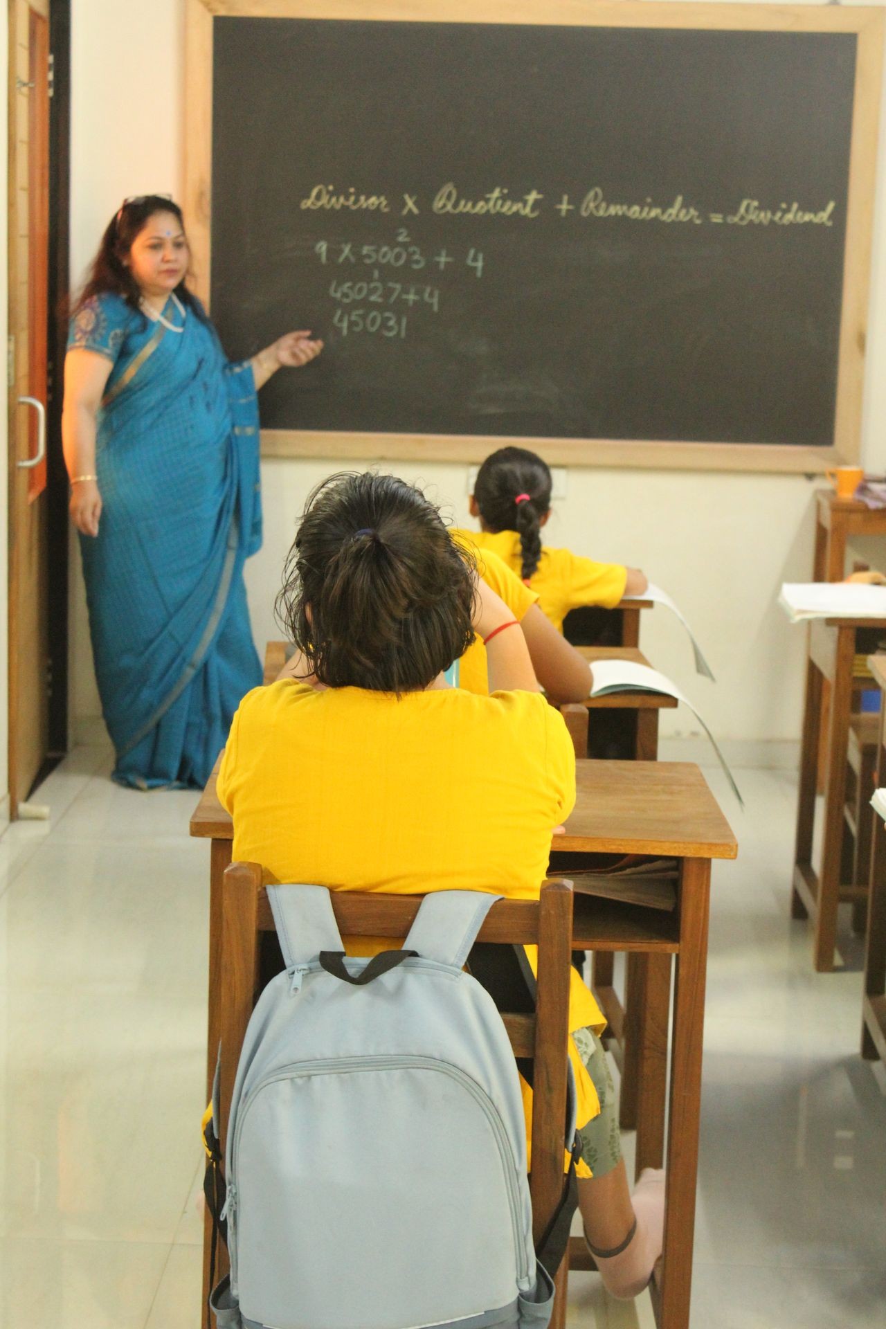 Teacher in blue saree explaining math on blackboard to students in yellow shirts in classroom.