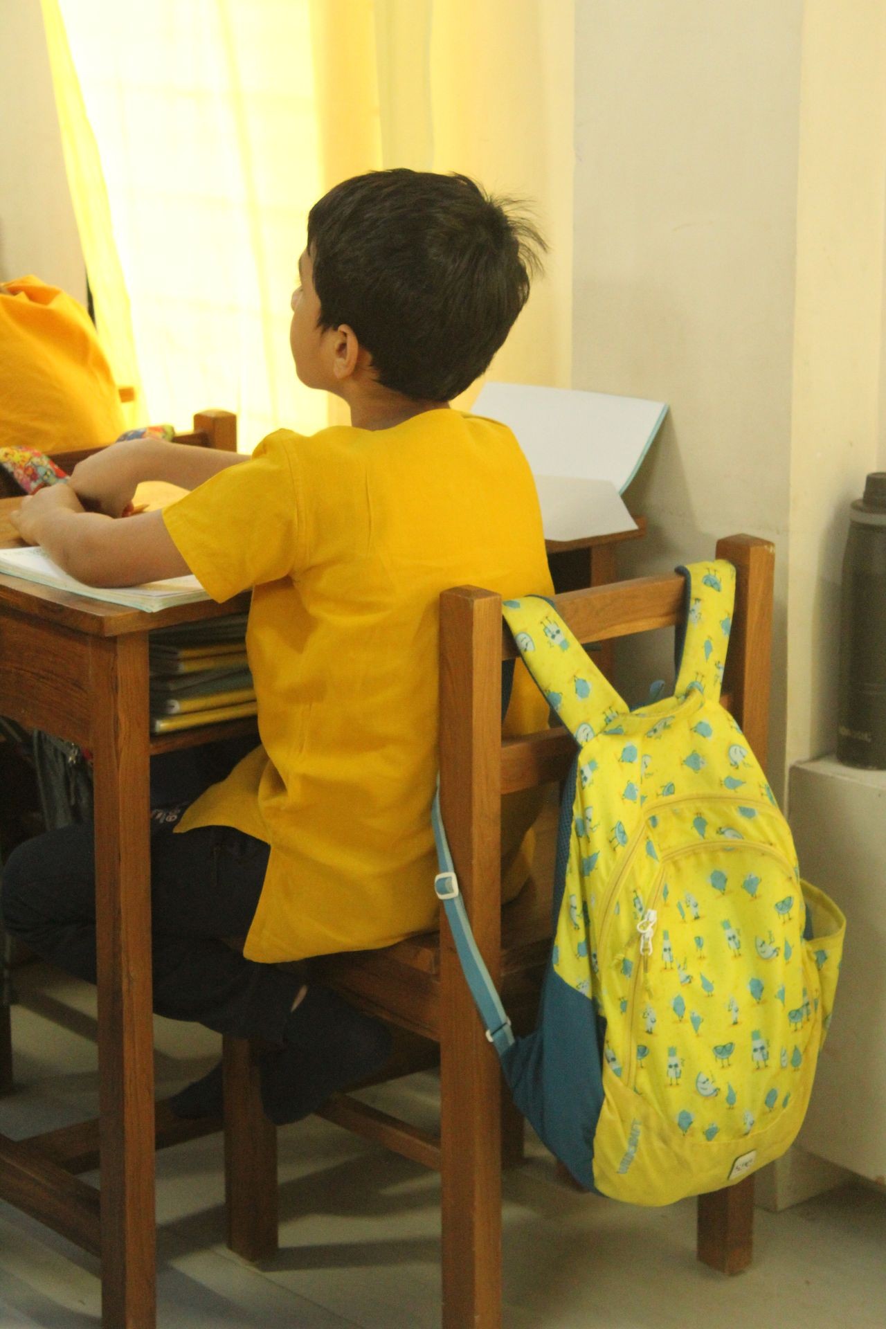 Child in yellow shirt sitting at desk with yellow backpack hanging on chair.
